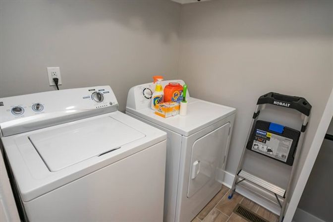 Compact laundry room with white top-load washing machine and matching front-load dryer, laundry detergent and supplies on dryer, Kobalt folding step ladder, gray walls and tile floor.