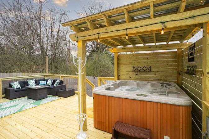 Cozy backyard wooden deck featuring a covered hot tub under a pergola with string lights, wicker patio seating with tropical cushions and a surrounding privacy fence