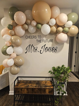 Neutral blush, olive, cream and tan balloon garland framing a bridal-shower backdrop on a modern home entryway wall, with a rustic wood bench and potted trailing plant.