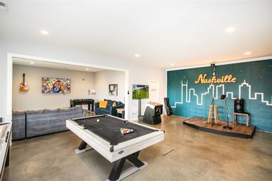 Bright Nashville game room with pool table, gray sectional lounge, wall-mounted TV and arcade machine, guitar on the wall, and a small performance stage under an illuminated skyline "Nashville" mural.