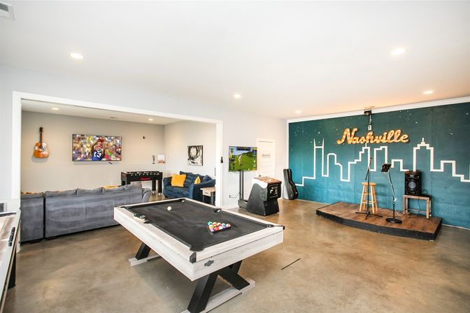 Bright Nashville game room with pool table, gray sectional lounge, wall-mounted TV and arcade machine, guitar on the wall, and a small performance stage under an illuminated skyline "Nashville" mural.