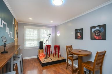 Cozy home music room with a small raised stage, microphone on a stand, two red bar stools and neon "Live Music" sign, bar counter seating, round wooden table, hardwood floors and whimsical cowboy-dog wall art by a bright window.