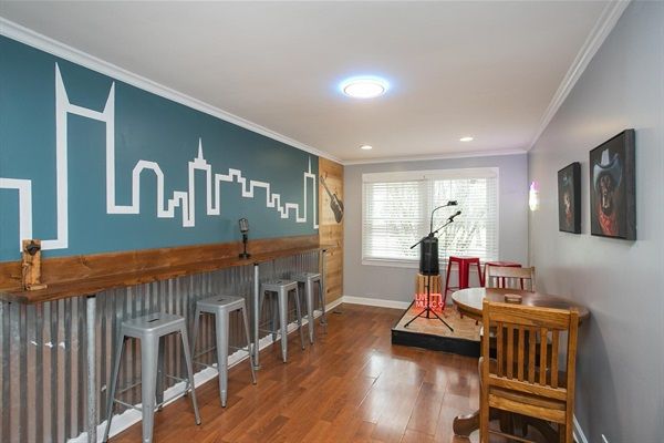 Home music room with teal city-skyline mural, corrugated metal bar and wood counter with metal stools, small stage with microphone, hardwood floors and window light