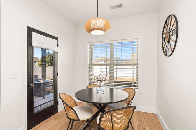 Cozy sunlit dining nook with round black table, four rattan chairs, woven pendant light, black glass door and window with blinds overlooking a suburban backyard, hardwood floors and wall clock.
