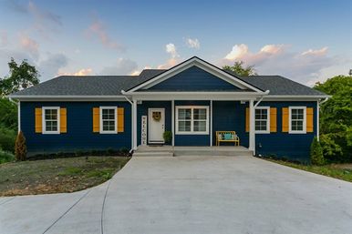 Front exterior of a single-story navy blue ranch-style suburban home with yellow shutters, covered porch with a Welcome sign and bench, wide concrete driveway and small landscaped yard at dusk.
