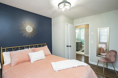Cozy modern bedroom with coral quilted bedding, navy accent wall and sunburst mirror, gold metal bedframe, velvet chair, wood floors and an open doorway to a bright ensuite bathroom.