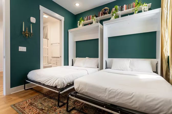 Cozy guest room with two white Murphy beds pulled down against an emerald green accent wall, crisp white linens, overhead bookshelf with books and trailing plants, patterned area rug, hardwood floor and an open en-suite bathroom.
