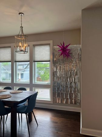Bright suburban dining room with a wooden table and blue chairs, modern glass pendant light, row of large windows, metallic silver foil backdrop and hanging pink star decoration