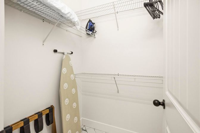 Bright minimalist walk-in closet in a modern home with white wire shelving, an ironing board and iron on the shelf, empty black hangers, a luggage rack, and marble-look tile floor.