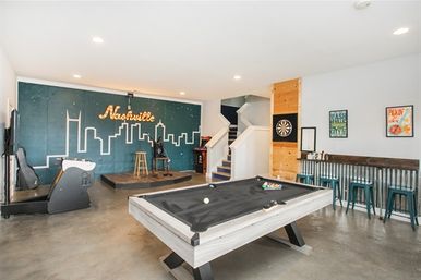 Bright Nashville game room featuring a gray pool table, small performance stage with illuminated skyline mural, dartboard and metal-bar seating