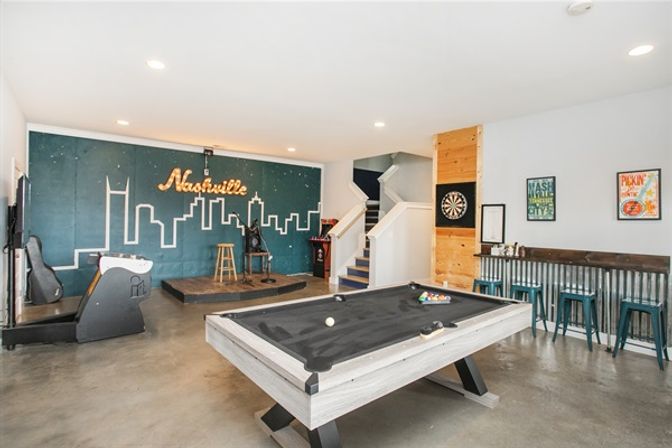 Bright Nashville game room featuring a gray pool table, small performance stage with illuminated skyline mural, dartboard and metal-bar seating