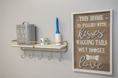Home entryway wall shelf with five hooks holding pet-care items—a gray tin canister, lint roller and small containers—beside a rustic wooden sign reading 'This home is filled with kisses, wagging tails, wet noses and love'.