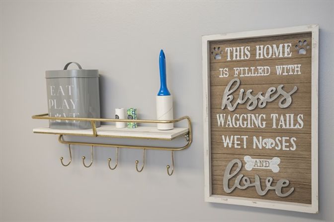 Home entryway wall shelf with five hooks holding pet-care items—a gray tin canister, lint roller and small containers—beside a rustic wooden sign reading 'This home is filled with kisses, wagging tails, wet noses and love'.
