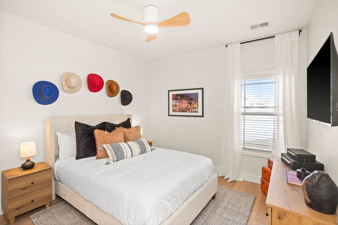 Modern cozy bedroom with white walls, queen bed layered with textured pillows, playful colorful hat display above the headboard, wooden nightstand and dresser, wall-mounted TV, ceiling fan, and sunlit window with sheer white curtains.