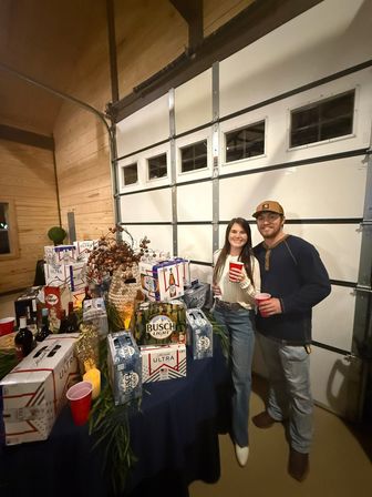 Couple smiling with red solo cups at a cozy wood‑paneled garage party beside a table stacked with beer cases, bottles and festive decorations.