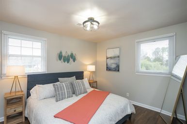 Cozy modern bedroom interior with navy upholstered headboard, white patterned bedding, coral throw, striped accent pillows, wooden nightstands with lamps, teal wall art and a leaning floor mirror.