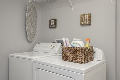 Cozy gray laundry room with white top-load washer and dryer, woven basket of detergent and spray on the dryer, a hanging ironing board, and decorative "Wash" and "Dry" wall signs.