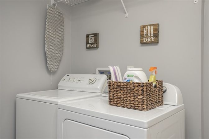 Cozy gray laundry room with white top-load washer and dryer, woven basket of detergent and spray on the dryer, a hanging ironing board, and decorative "Wash" and "Dry" wall signs.