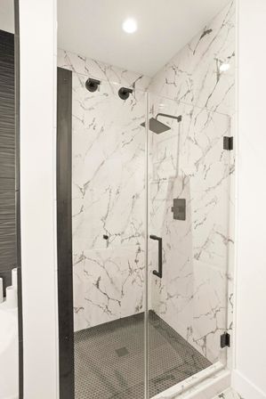 Sleek modern walk-in bathroom shower with frameless glass door, white marble-look walls with gray veining, matte black rain showerhead and hardware, and dark hexagonal mosaic tile floor.