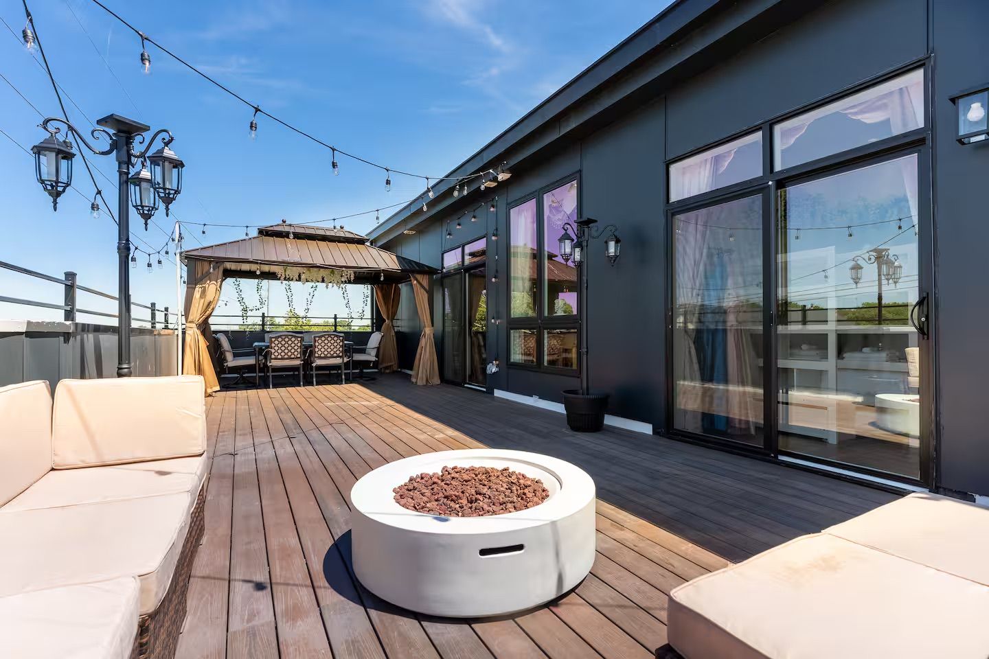Sunlit rooftop deck with wood planks, cushioned beige lounges, round modern fire pit, curtained gazebo dining area, string lights and decorative lanterns under a bright blue sky