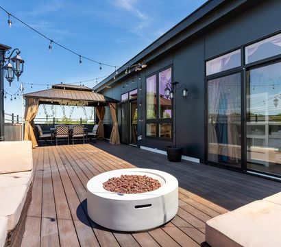 Sunlit rooftop deck with wood planks, cushioned beige lounges, round modern fire pit, curtained gazebo dining area, string lights and decorative lanterns under a bright blue sky