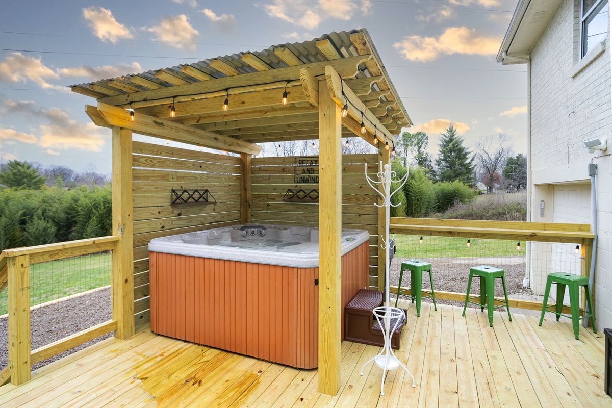Backyard wooden deck with covered pergola sheltering an orange-sided hot tub, string lights, green metal stools, and a grassy suburban yard at sunset.
