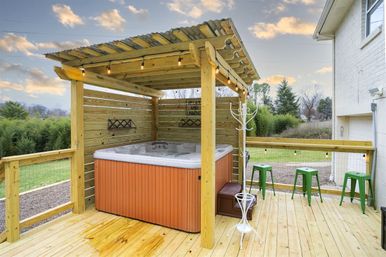 Backyard wooden deck with covered pergola sheltering an orange-sided hot tub, string lights, green metal stools, and a grassy suburban yard at sunset.