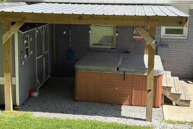 Backyard covered hot tub tucked under a wooden pergola next to a gray storage shed, sitting on a gravel pad with wooden steps up to a brick house window.
