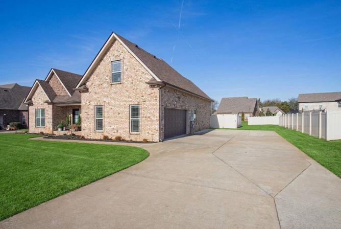 Sunny suburban brick home with attached two-car garage, wide concrete driveway, neatly trimmed green lawn and white privacy fence under a clear blue sky.