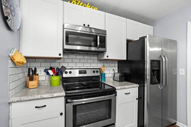 Sleek modern kitchen with white cabinets, subway-tile backsplash and speckled granite countertop, stainless steel microwave, electric range and side-by-side refrigerator, plus a knife block, utensil holder and toaster on the counter.