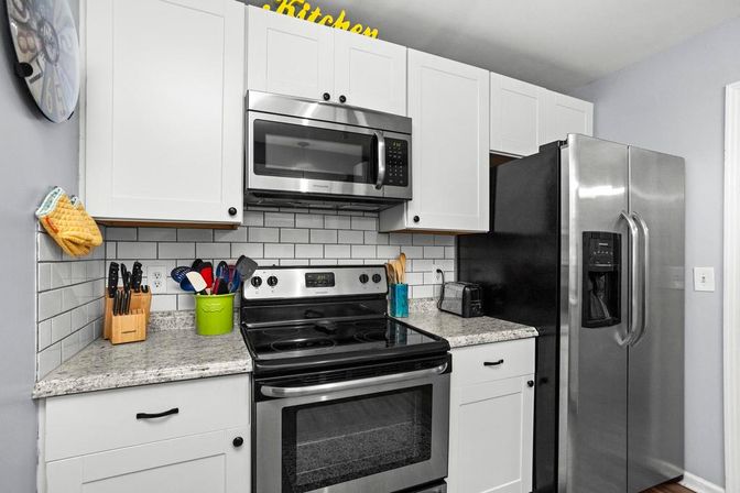 Sleek modern kitchen with white cabinets, subway-tile backsplash and speckled granite countertop, stainless steel microwave, electric range and side-by-side refrigerator, plus a knife block, utensil holder and toaster on the counter.