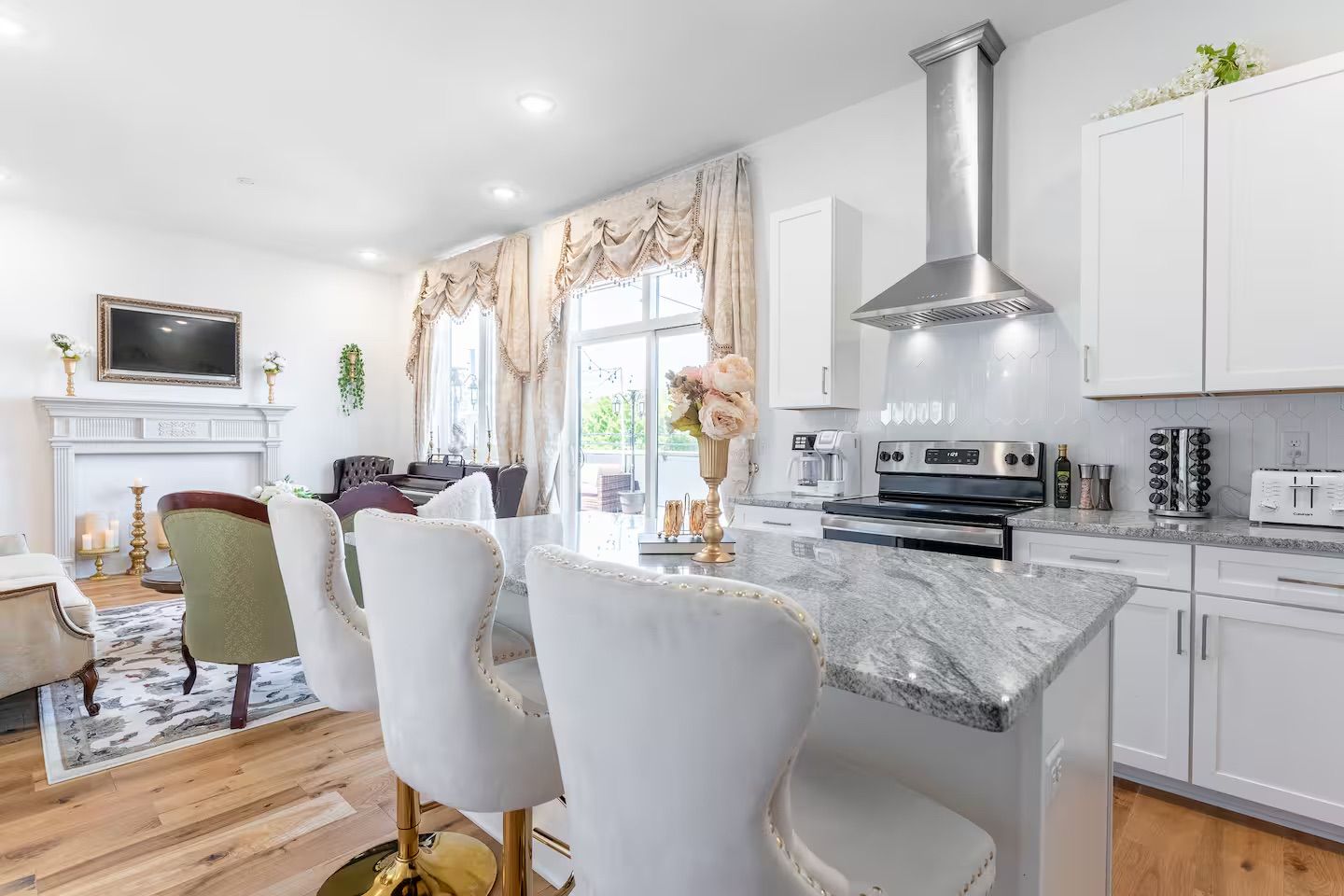 Bright, elegant open-concept kitchen and living room with marble island and gold-based tufted bar stools, stainless steel range hood, white cabinetry, hardwood floors, ornate French-style drapes, and a decorative fireplace.
