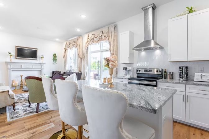 Bright, elegant open-concept kitchen and living room with marble island and gold-based tufted bar stools, stainless steel range hood, white cabinetry, hardwood floors, ornate French-style drapes, and a decorative fireplace.
