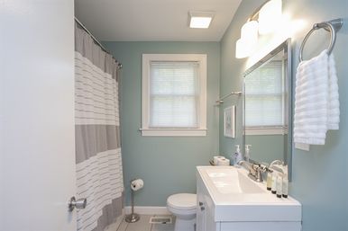 Cozy residential bathroom with seafoam-green walls, white vanity and sink, large mirror with lights, striped gray-and-white shower curtain, toilet beneath a window with blinds, and a white towel on a ring.