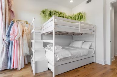 Cozy white wooden bunk bed in a bright kids' bedroom with a lower full bed, built-in stairs, folded towels, pastel dresses on a gold clothing rack, hardwood floors and hanging green vines