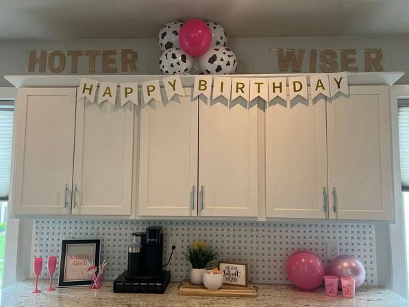 Home kitchen birthday display with modern white cabinets and mosaic tile backsplash, a 'HAPPY BIRTHDAY' banner and gold glitter 'HOTTER' and 'WISER' letters, pink and spotted balloons, coffee maker and pink cups on a granite countertop.