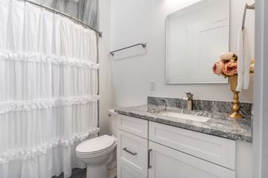 Bright modern white bathroom with ruffled white shower curtain, compact toilet, white shaker vanity topped with gray granite countertop and undermount sink, brushed nickel faucet, framed mirror, dark hex-tile floor, and gold vase of pink peonies.
