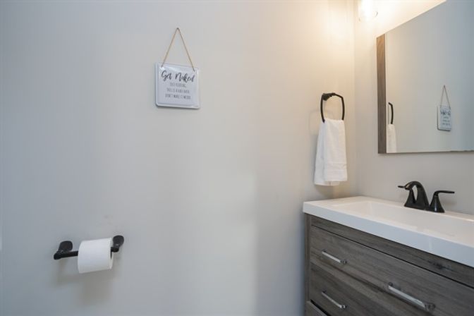 Sleek modern powder room with gray wood vanity, white countertop and black fixtures, towel ring with white towel, toilet paper holder, mirror and small hanging sign.