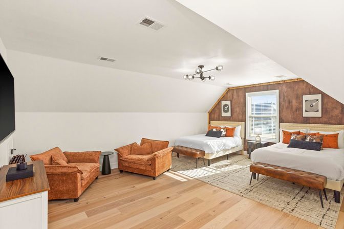 Sunlit attic loft bedroom with two beds, tufted leather benches at the foot of each bed, rustic wood accent wall, matching orange velvet lounge chairs, modern sputnik chandelier and light hardwood floors.