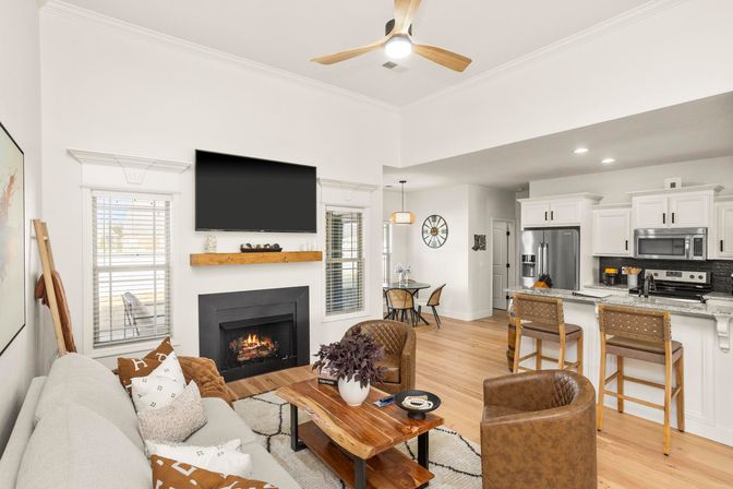 Bright modern open-concept living room and kitchen with a lit fireplace beneath a wall-mounted TV, wood coffee table, leather accent chairs, breakfast bar with stools, white cabinets and stainless appliances.