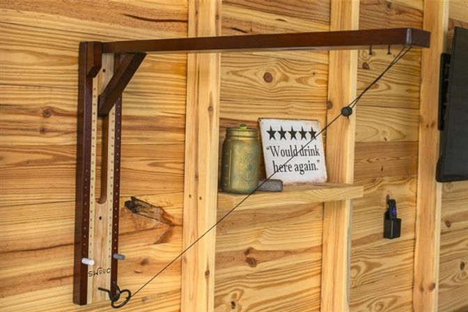 Rustic pine-plank shed wall with an adjustable wooden arm and pulley cable, small shelf holding a jar and a five-star sign reading 'Would drink here again'.