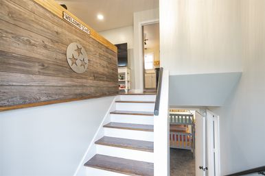 Light-filled modern home interior: white staircase with dark wood treads leading to an open landing beside a reclaimed wood accent wall with circular three-star metal decor and a small “Welcome” sign; kitchen doorway ahead and bunk beds visible below.