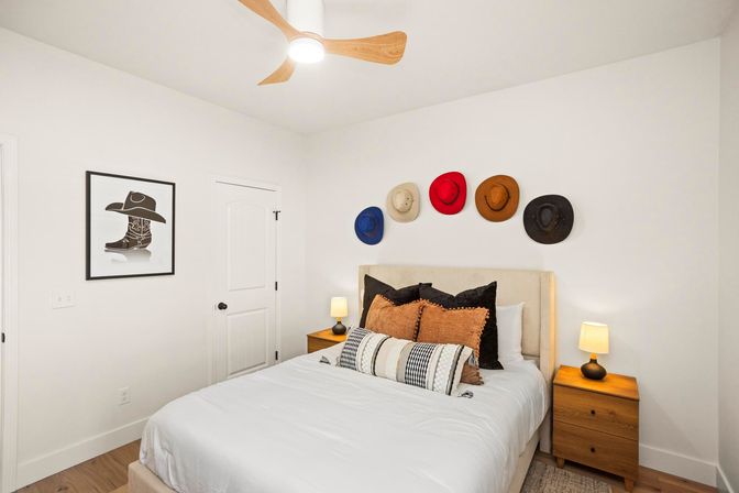 Bright modern bedroom with white walls and ceiling fan, queen bed with textured headboard and layered pillows, two wooden nightstands with lamps, and a playful row of colorful cowboy hats mounted above the bed.