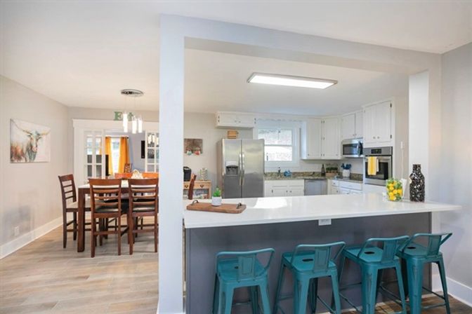 Open-concept modern kitchen and dining area in a suburban home with white cabinets, stainless steel appliances, granite counters, a long white breakfast bar with four teal metal stools, and a wooden dining table under pendant lights