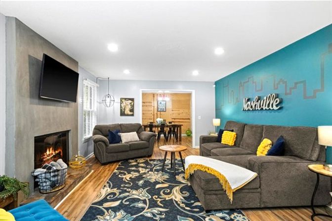 Cozy, modern living room with a lit fireplace and wall-mounted TV, teal accent wall featuring a Nashville skyline sign, gray sectional sofas with yellow throw pillows, patterned area rug and hardwood floors.