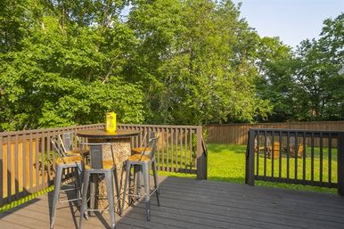 Sunny backyard deck with round outdoor bar table topped by a yellow lantern, four metal bar stools, dark wooden railing, green lawn, tall leafy trees and a wooden fence