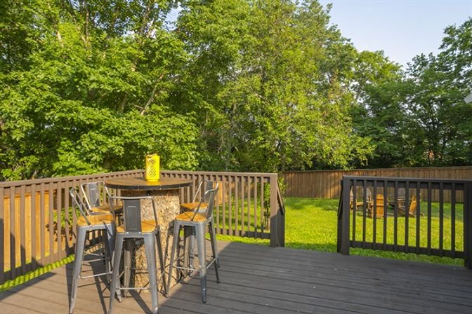 Sunny backyard deck with round outdoor bar table topped by a yellow lantern, four metal bar stools, dark wooden railing, green lawn, tall leafy trees and a wooden fence