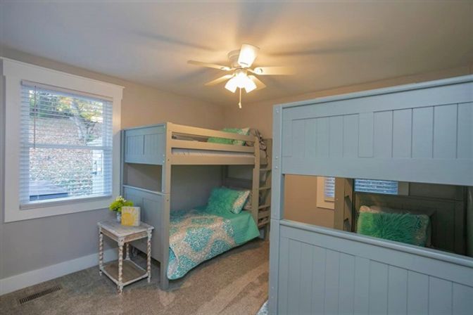 Cozy bedroom with gray wooden bunk beds, teal patterned bedding and fluffy pillows, white ceiling fan, window with blinds, distressed nightstand and carpeted floor