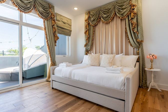 Sunlit luxury bedroom with white daybed, plush pillows and folded towels, ornate green-and-gold drapery, sliding glass door to a balcony lounge, and warm hardwood floors with a small floral side table.