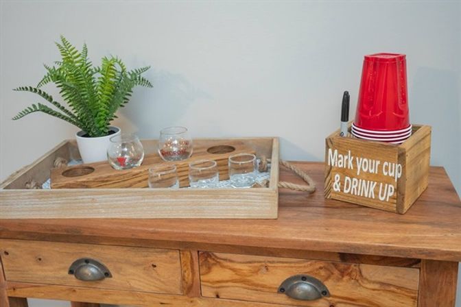 Home party setup on a wooden console: rustic tray with potted fern and shot glasses, beside a wooden box holding stacked red plastic cups and a marker with the sign 'Mark your cup & DRINK UP'.